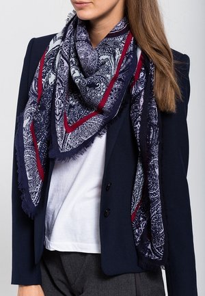 Femme portant un blazer bleu marine et un haut blanc, accessoirisée d'une grande écharpe à motifs bleu marine, blanc et rouge drapée autour du cou.