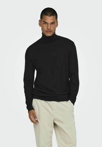 Zwarte coltrui van zachte stof, met lange mouwen en een geribde onderrand, gecombineerd met lichtbeige, gestructureerde broek.