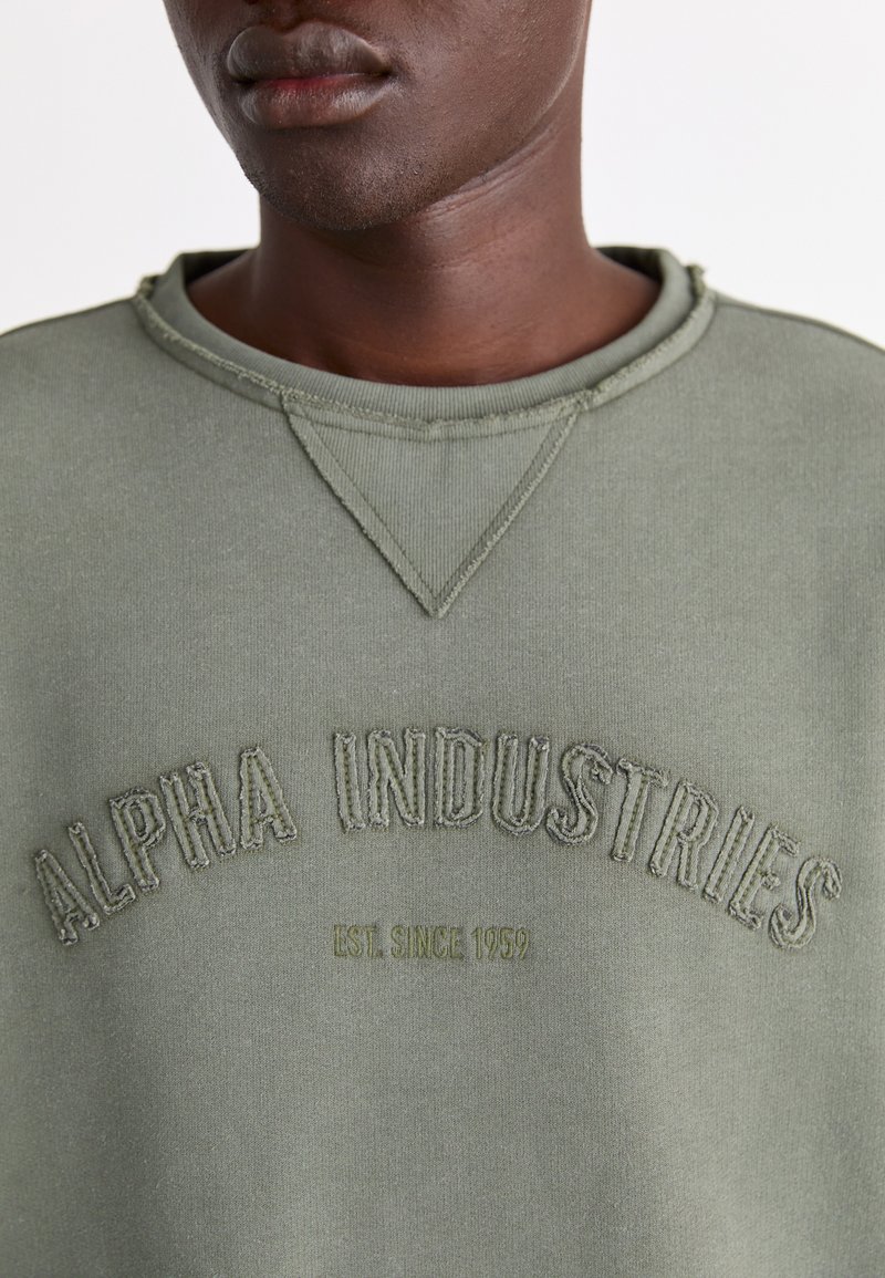 Olivgrüner Sweatshirt mit Rundhalsausschnitt, strukturiertem Nähen und erhabener Stickerei "ALPHA INDUSTRIES EST. SINCE 1959" auf der Vorderseite.