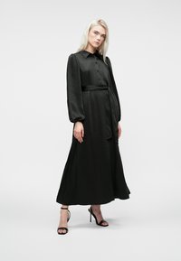 Mustas pikkade varrukatega kleit, mis on valmistatud siledast kangast, nööpidega esiosa, krae ja vöödiga talje juures. Omab voogavat seelikut ja stiilseid musti kontsseid kingi.