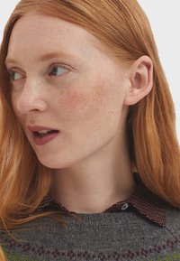 Pull en laine gris avec un motif multicolore au bord, porté sur une chemise à col rouge foncé avec des rayures blanches. Cheveux auburn clair, peau tachetée de rousseurs.