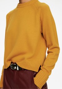 Senfgelber Strickpullover mit geripptem Rundhalsausschnitt und Bündchen. Hat eine leicht lockere Passform und seitliche Schlitze. Kombiniert mit bordeauxroten Hosen und einem Gürtel.