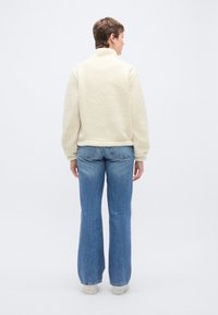 Jakna od flisa krem boje s visokim ovratnikom, uparena s plavim trapezasto krojenim trapericama i bijelim tenisicama. Jakna ima tkaninu s teksturom.