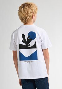 Bílý bavlněný tričko s grafickým designem na zádech, zahrnujícím černé a modré tvary a abstraktní motiv listu.