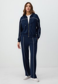 Navy tracksuit with white piping; zip-up hooded jacket and straight-leg pants; soft fabric; features side pockets and white sneakers.