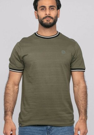 Homme barbu portant un t-shirt côtelé vert olive avec un col et des bords de manches rayés noirs et blancs, associé à un jean bleu.