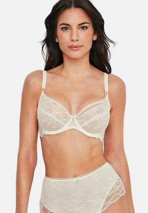 Femme portant un soutien-gorge en dentelle crème et une culotte taille haute en dentelle assortie, debout devant un fond blanc uni.