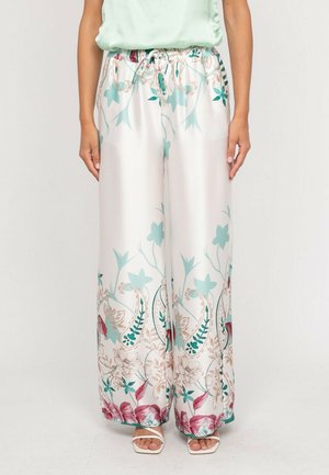 Pantalon large en satin avec motifs floraux turquoise, beige et rose, taille avec cordon de serrage, porté avec des sandales blanches à bouts ouverts.
