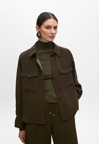 Camicia marrone oversize con due tasche sul petto, chiusura a bottoni e dettagli in tonalità oro. Indossata sopra un dolcevita verde coordinato e pantaloni.
