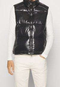 Schwarze Steppweste mit hohem Kragen, glänzender Oberfläche, Reißverschluss vorne und großen gepolsterten Abschnitten, die über einem weißen Langarmshirt getragen wird.