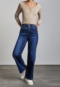 Donna che indossa un top henley beige a maniche lunghe, jeans blu scuro a vita alta e sneaker bianche, in piedi con le mani in tasca.
