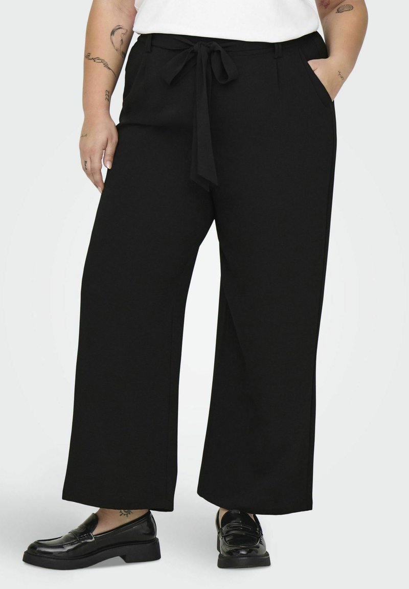Pantalones de pierna ancha negros, fabricados en un tejido suave, con una cinturilla atada y bolsillos frontales. Combinados con unos mocasines negros brillantes.
