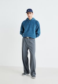 Felpa blu in pile con tasca a marsupio, pantaloni grigi a gamba larga e sneakers grigi, abbinati a un cappellino blu navy con un logo.