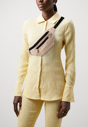 Riñonera a rayas beige llevada en el hombro, con dos compartimentos con cremallera, combinada con una blusa de textura ligera en color amarillo y pantalones.