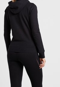 Sudadera negra con capucha forrada, puños y dobladillo acanalados. Combinada con leggings negros con cortes laterales para ventilación. Textura de tela suave.