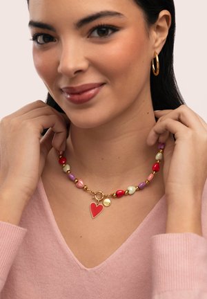 Donna che indossa un maglione rosa e una collana colorata di perline con un ciondolo a forma di cuore rosso, che tiene la collana vicino al collo e sorride dolcemente.