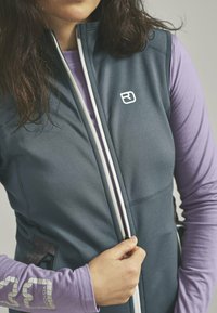 Dunkelgraue ärmellose Weste mit einem weißen Reißverschluss und Logo, getragen über einem helllila Langarmshirt; verfügt über seitliche Taschen.