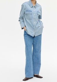 Camicia di denim azzurro chiaro con colletto, chiusura a bottoni e due tasche sul petto, abbinata a pantaloni a gamba larga coordinati e scarpe marroni scure.