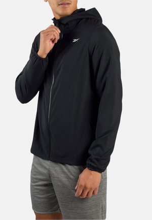 Uomo che indossa una giacca nera con cappuccio e cerniera e pantaloncini sportivi grigi, mentre sistema la cerniera della giacca.