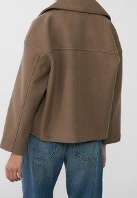 Brown wool blend jacket with a wide collar, dropped shoulders, and a boxy shape, paired with light blue denim jeans.