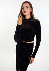 Set din catifea neagră în două piese, compus dintr-un top scurt cu mâneci lungi și embellishe pe umeri, și o fustă asortată cu o fanta pe o parte și accente cu butoane.