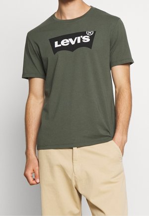 Homme portant un t-shirt Levi's vert olive et un pantalon beige, debout avec une main dans la poche devant un fond uni.