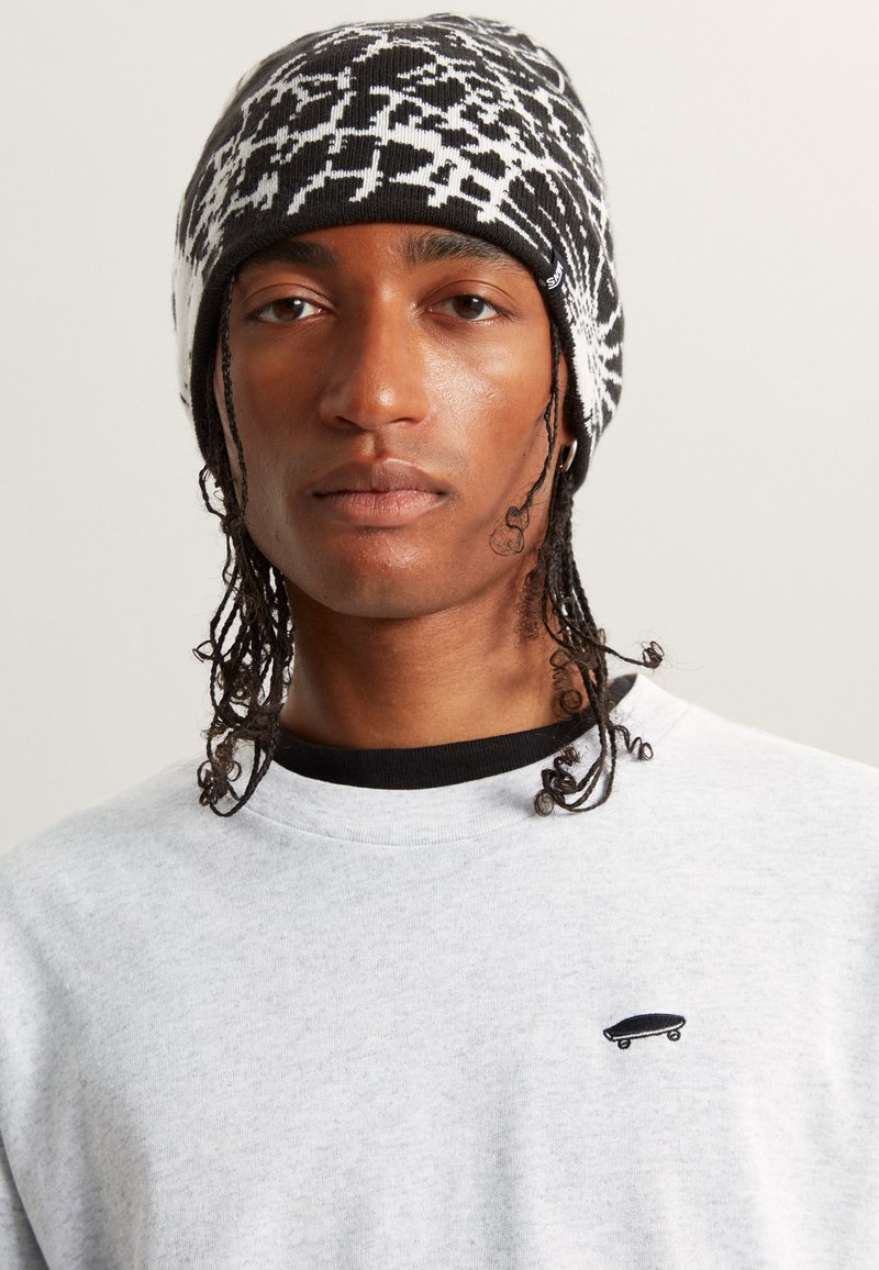 Berretto lavorato a maglia con un motivo geometrico in bianco e nero, abbinato a una felpa grigia chiara con un piccolo logo di skateboard nero.