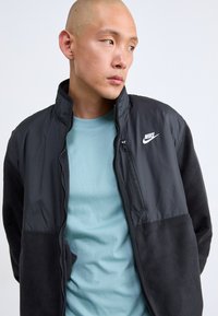 Moški z obrito glavo nosi črno jakno Nike preko svetlo modre srajce, gleda na stran proti enostavni svetli ozadju.