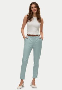 Pantaloni azzurri su misura con risvolti, tasche frontali e una cintura marrone, abbinati a un top bianco aderente.