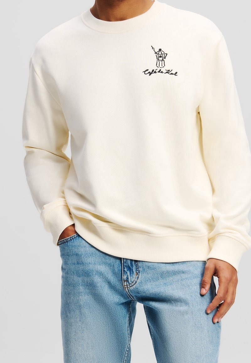 Homme portant un sweat-shirt crème avec une petite cafetière noire et le texte « Cafe de Kol » sur le côté gauche de la poitrine, associé à un jean bleu clair.