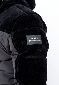 Svart vadderad jacka med en texturerad, plyschliknande tyg och en slät nylonfinish. En rektangulär etikett på ärmen har texten "LA ROSA COPENHAGEN."