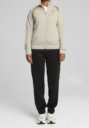 Personne portant une veste beige zippée avec des empiècements marron aux épaules, pantalon noir et baskets blanches, debout devant un fond clair uni.