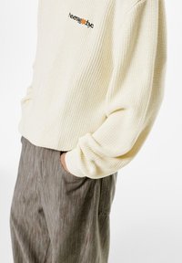 Pulover ribbed de un bej deschis cu un mic logo brodat în negru și portocaliu, asociat cu pantaloni gri cu dungi verticale. Detalii ale texturii materialului vizibile.