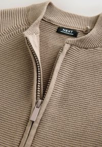 Maglione beige con zip e texture a coste, caratterizzato da un colletto alto e sottili righe verticali, completato da una cerniera leggera.