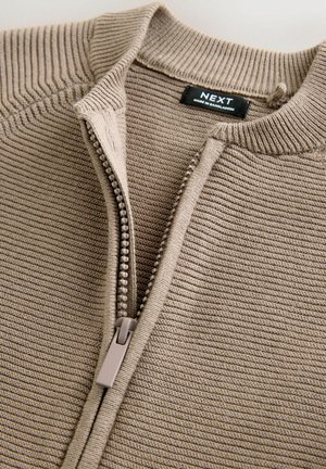 Beżowy sweter na zamek z prążkowaną fakturą, z kołnierzem typu stójka i delikatnymi pionowymi paskami, uzupełniony lekkim zamkiem błyskawicznym.