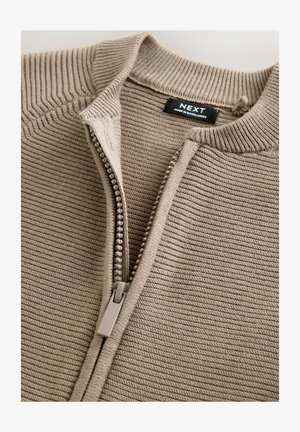 Bež pulover na zadrgo z rebrasto teksturo, z ovratnikom stoječega kroja in subtilnimi navpičnimi črtami, dopolnjen z lahkotno zadrgo.