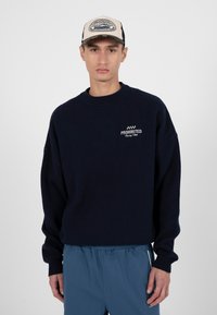 Jeune homme portant un sweat noir "Prohibited Racing Club", un pantalon bleu et une casquette beige et noire avec le même logo sur fond blanc.