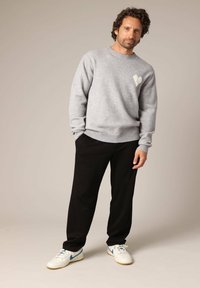 Pull en maille gris clair avec un motif cœur, associé à un pantalon noir et des baskets blanches avec des accents bleus. Le mannequin est en pose neutre.