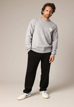 Lichtgrijze gebreide trui met een hartdetail, gecombineerd met zwarte broek en witte sneakers met blauwe accenten. Het model staat in een neutrale pose.