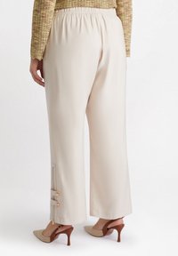Szerokie, kremowe spodnie z elastycznym paskiem w talii, z zamkami błyskawicznymi i złotymi akcentami na mankietach, zestawione z beżowymi szpilkami.