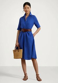 Niebieska lniana sukienka koszulowa z krótkimi rękawami, z paskiem w talii. Połączona ze słomkową torebką i brązowymi sandałami na paski.