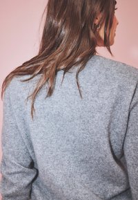 Pull en laine gris avec une finition texturée, affichant une coupe décontractée et des épaules tombantes, vu de l'arrière sur un fond rose.