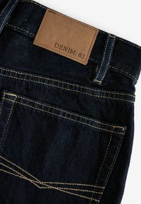 Tumedat denim püksid pruuni nahaplaadi sildiga "DENIM: 82." Omavad kontrastset õmblust ja ühte tagumist taskut dekoratiivsete õmblustega.
