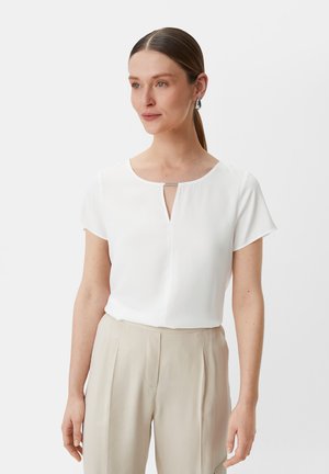 White short-sleeve blouse featuring a keyhole neckline with a silver accent, paired with light beige trousers. Smooth fabric texture.