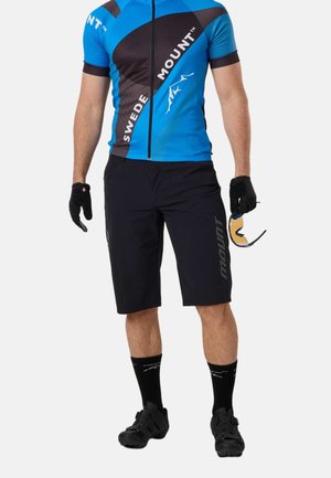 Mand iført blå og sort cykeltrøje og sorte shorts, der holder solbriller, med sorte handsker, sokker og cykelsko.