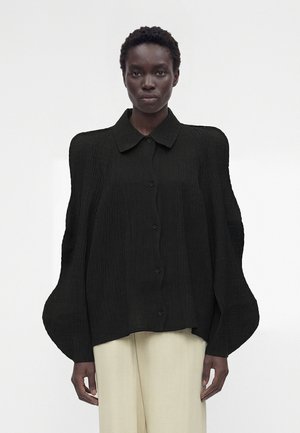 Kobieta o ciemnej karnacji, ubrana w czarną teksturowaną koszulę z dużymi bufkami na rękawach oraz beżowe spodnie na jednolitym białym tle.