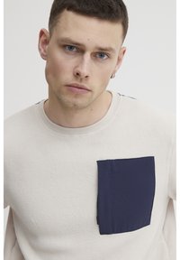 Sudadera de forro polar beige con un bolsillo azul marino, escote redondo y ligera textura. Cuenta con una cremallera en el bolsillo para mayor utilidad.