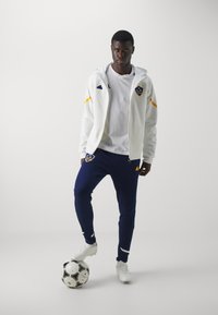 Witte zip-up jas met gele accenten, gecombineerd met marineblauwe broek en witte voetbalschoenen. Houdt een zwart-wit voetbals voorzien.