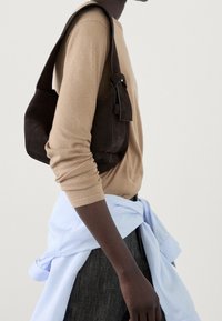 Personne à la peau foncée portant une chemise beige à manches longues et un pantalon foncé, portant un sac à bandoulière en daim foncé avec une sangle et un détail de boucle.