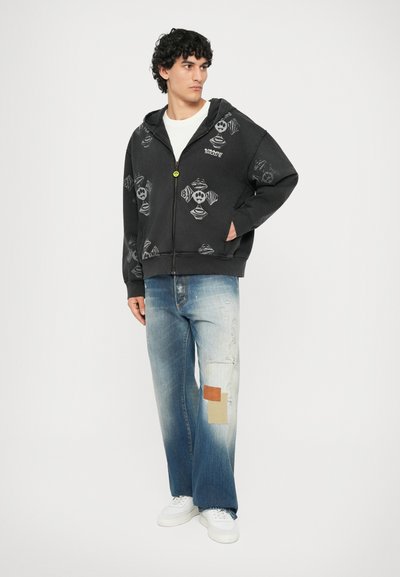 Sort zip-up hættetrøje med grafiktryk, der har en frontlomme og en rund gul lynlås. Blå slidte vide jeans med lapper.